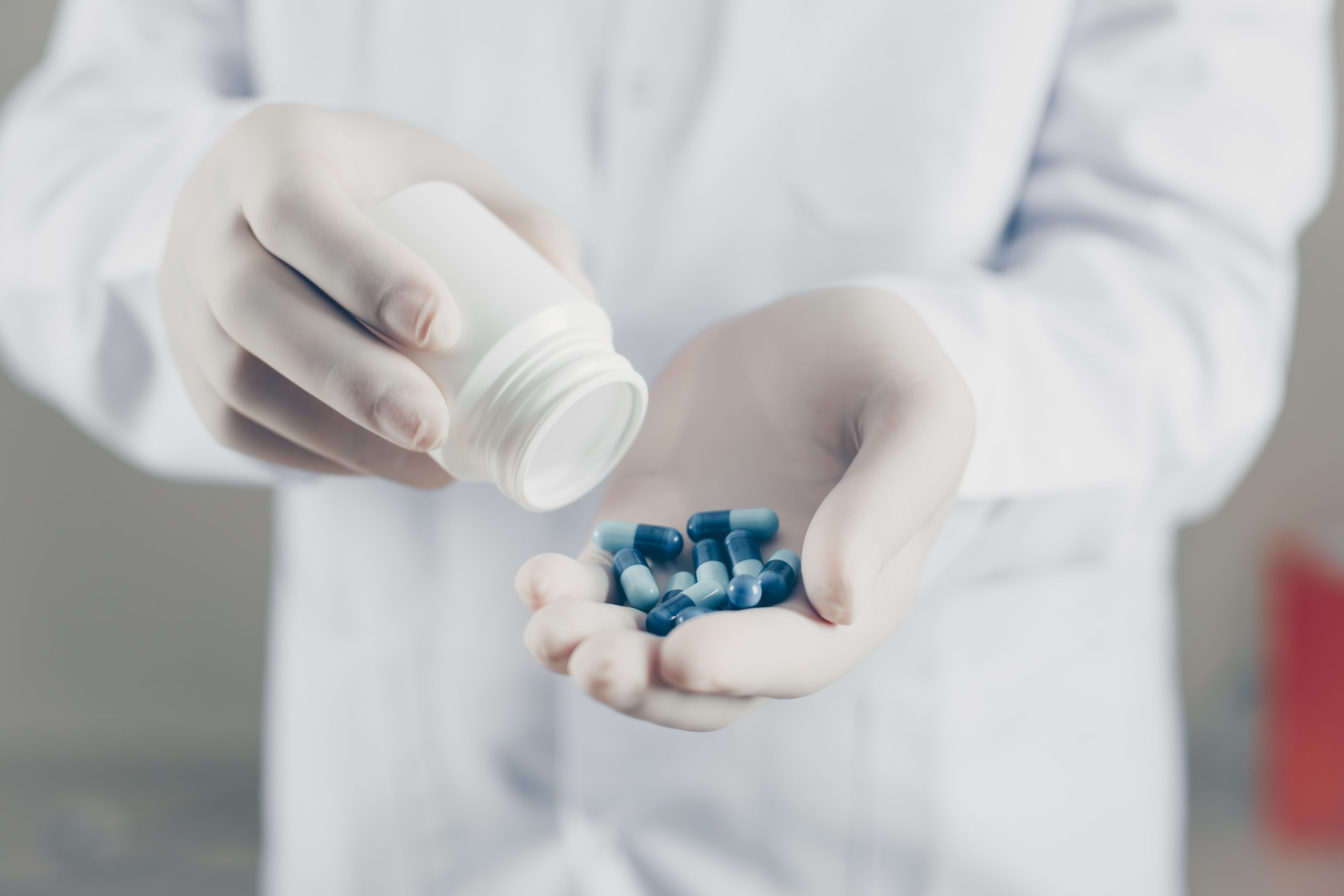 Doctor with gloves holding pills and bottle on gray background, side view.